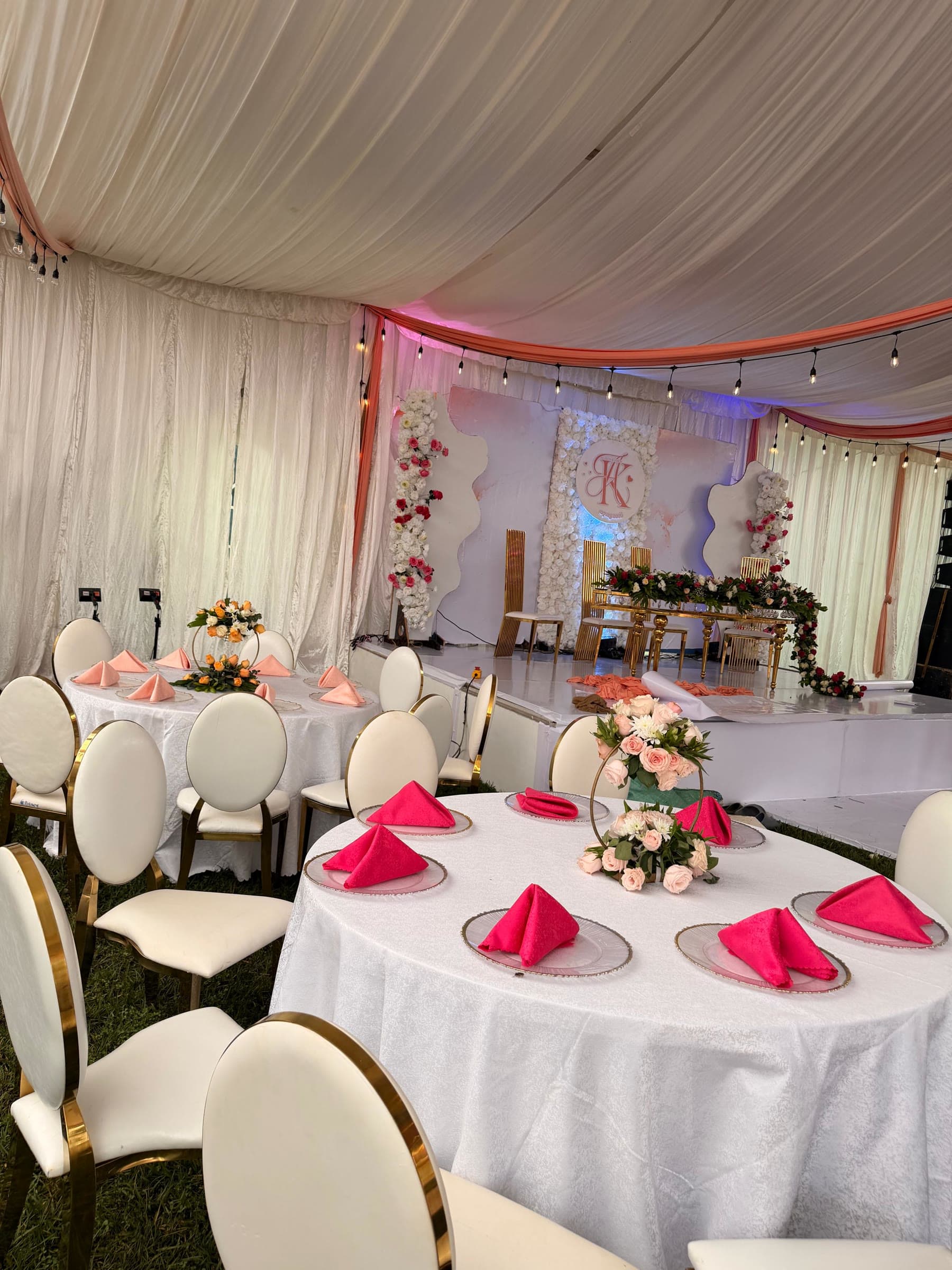 Head table on a raised stage beneath a monogrammed flower wall, with gilt chairs and a garland of red and white roses running the length of the table